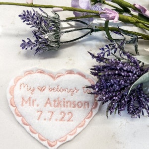 May include: White heart-shaped fabric patch with pink embroidered lettering that reads "My heart belongs to Mr. Atkinson 7.7.22". The patch is surrounded by a pink scalloped border. The patch is lying on a white surface with purple flowers.