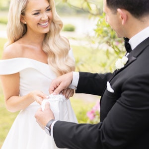 May include: A bride in a white off-the-shoulder wedding dress smiles at the groom, who is adjusting the lace on her dress.