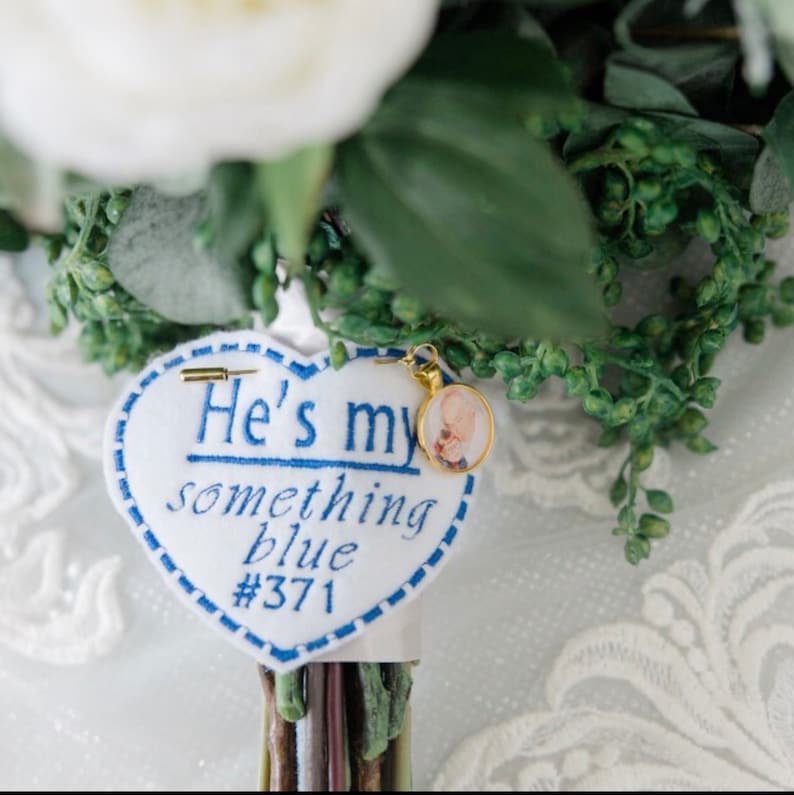 May include: A white heart-shaped fabric patch with blue embroidery that reads "He's my something blue #371". A small gold charm with a photo of a baby is attached to the patch.