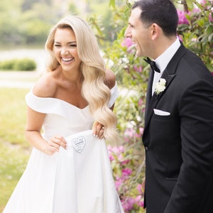 May include: A bride and groom laughing together outdoors. The bride is wearing a white off-the-shoulder wedding dress and holding up the skirt to reveal a heart with the words "We're finally married" written inside. The groom is wearing a black tuxedo with a white shirt and a black bow tie.