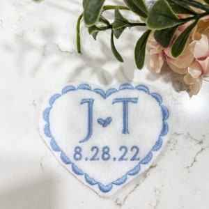 May include: A white heart-shaped patch with blue scalloped edges. The patch is embroidered with the initials "J & T" and the date "8.28.22".