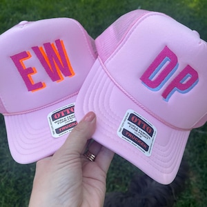 May include: Two pink trucker hats with the letters "EW" and "DP" embroidered on the front. The "EW" hat has orange and pink lettering, while the "DP" hat has blue and pink lettering. Both hats have a mesh back and a snap closure.