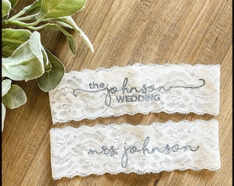 May include: Two white lace garters with gray embroidery. The top garter says "the johnson wedding" and the bottom garter says "mrs johnson".