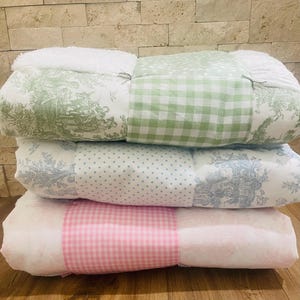 May include: A stack of four patchwork quilts in various patterns and colors. The top quilt is green and white gingham, the second is blue and white polka dot, the third is pink and white gingham, and the bottom is white with a green and white floral pattern.