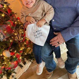 May include: A young child is holding a white cloth with black embroidery that reads "We ♥ Papa! Joey, Ashley,  Mila & Ellie". The child is wearing a tan hoodie, blue jeans, and pink shoes. The child is sitting on the lap of an adult wearing a blue button-down shirt and blue jeans.