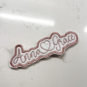 May include: A white felt patch with a pink embroidered design. The design says "Anna Grace" with a heart in between the names.