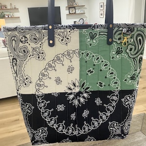 May include: A quilted tote bag featuring a patchwork design of black, white, and sage green paisley bandana patterns. The bag has two dark blue leather-like straps attached with gold-tone rivets and includes a gold-tone metal lobster clasp.
