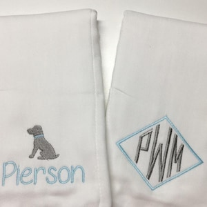 May include: Two white burp cloths with embroidered designs. One cloth features a gray dog silhouette with the name "Pierson" embroidered below. The other cloth has a diamond-shaped monogram with the initials "P.W.M."