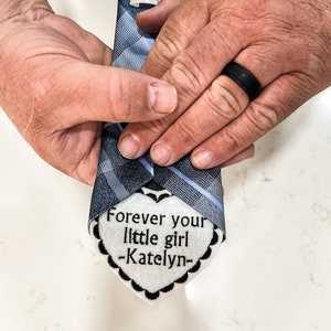 May include: A blue and white patterned tie with a white heart-shaped tag attached. The tag has the text "Forever your little girl -Katelyn-."