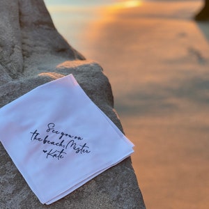 May include: White fabric square with black text that reads "See you on the beach! Mister Kate" with a small heart symbol before the name.