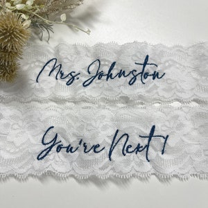 May include: Two white lace garters with blue embroidery. The top garter says "Mrs. Johnston" and the bottom garter says "You're Next".