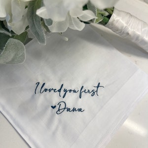 May include: White handkerchief with embroidered text "I loved you first Dana" in navy blue. A small navy blue heart is embroidered before the name. A white floral bouquet is in the background.