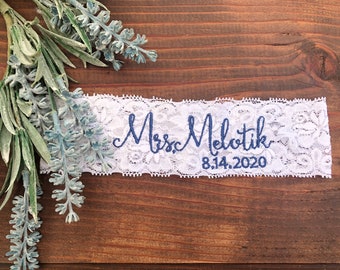 May include: White lace garter with blue embroidery that reads "Mrs. Melotnik 8.14.2020".