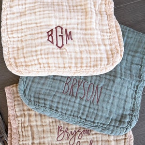 May include: Three muslin baby burp cloths with embroidered names. The top cloth is white with the monogram "BGM" in maroon thread. The middle cloth is green with the name "Bryson" embroidered in maroon thread. The bottom cloth is beige with the name "Bryson Michael" embroidered in maroon thread.