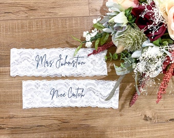 May include: Two white lace garters with blue embroidery. The top garter says "Mrs. Johnston" and the bottom garter says "Nice Catch".
