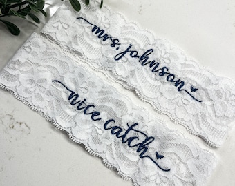 May include: Two white lace garters with blue embroidery. The top garter says "mrs. johnson" and the bottom garter says "nice catch".