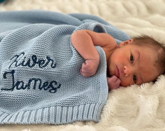 May include: A light blue knitted baby blanket with the name "River James" embroidered in dark blue thread. A newborn baby is lying on the blanket.