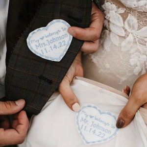 May include: Two white heart-shaped patches with blue embroidery. The text on the patches reads "My heart belongs to Mrs. Johnson 11.14.22" and "My heart belongs to Mr. Johnson 11.14.22".