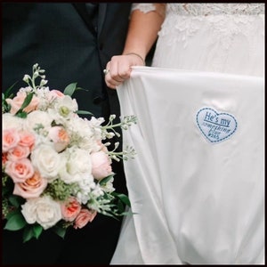 May include: A bride holds up the bottom of her white wedding dress, revealing a blue heart-shaped patch that says "He's my something blue #265".