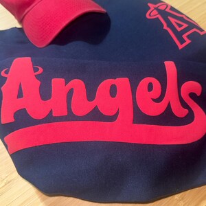 May include: A red baseball cap with the letter "A" and a navy blue sweatshirt with the word "Angels" in red. The sweatshirt also features a red "A" logo. The items are likely sports apparel.