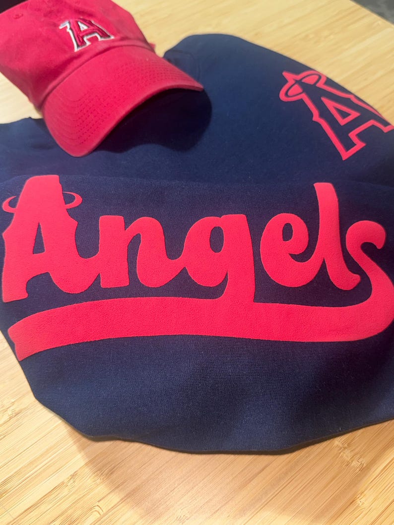 May include: A red baseball cap with the letter "A" and a navy blue sweatshirt with the word "Angels" in red. The sweatshirt also features a red "A" logo. The items are likely sports apparel.