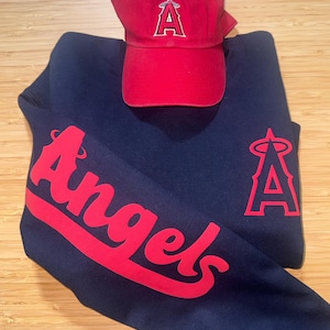 May include: A navy blue sweatshirt and a red baseball cap. The sweatshirt features the word "Angels" in red script on the sleeve and a red "A" logo. The red cap has a matching "A" logo.