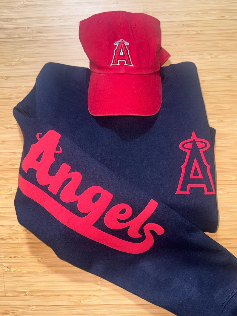 May include: A navy blue sweatshirt and a red baseball cap. The sweatshirt features the word "Angels" in red script on the sleeve and a red "A" logo. The red cap has a matching "A" logo.