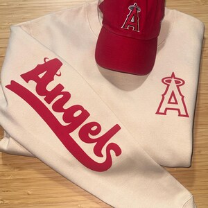 May include: A cream-colored sweatshirt and a red baseball cap. The sweatshirt features the word "Angels" in red script on the sleeve and a red "A" logo on the chest. The red cap has the same "A" logo.