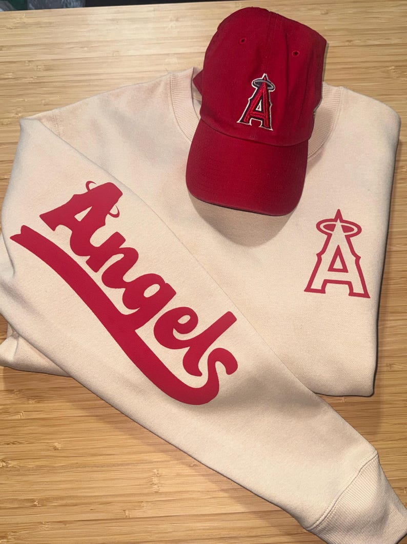 May include: A cream-colored sweatshirt and a red baseball cap. The sweatshirt features the word "Angels" in red script on the sleeve and a red "A" logo on the chest. The red cap has the same "A" logo.