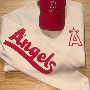 May include: Cream-colored sweatshirt and red baseball cap. The sweatshirt features the word "Angels" in red script on the sleeve and a red "A" logo. The red cap also has the "A" logo.