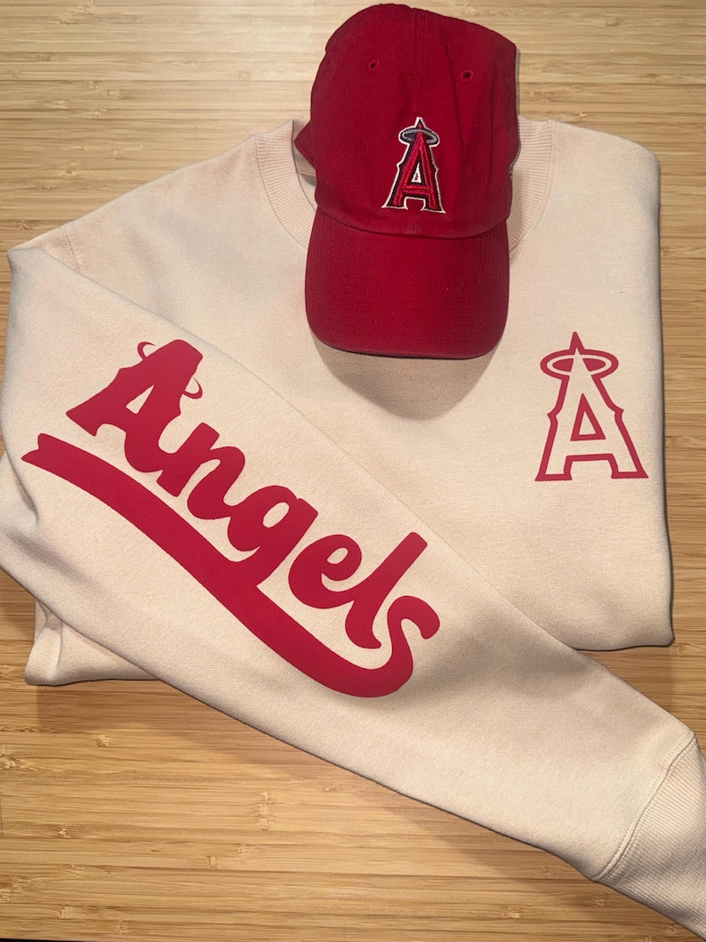 May include: Cream-colored sweatshirt and red baseball cap. The sweatshirt features the word "Angels" in red script on the sleeve and a red "A" logo. The red cap also has the "A" logo.