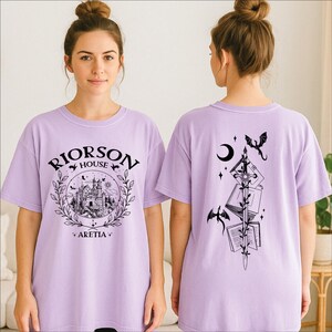 A woman wearing a purple t-shirt with a graphic design on the front and a back featuring a black and white illustration of a castle, a crescent moon, and a dragon.