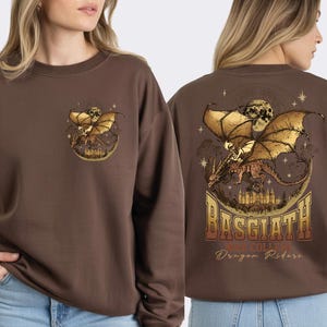 a woman wearing a brown sweater with a graphic design featuring a dragon and a castle. The design includes the text "BASGLATH WAR COLLEGE Dragon Riders" on the back of the sweater.