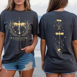 two women wearing black t-shirts with gold designs on them. The t-shirts have a dragon and star motif, and the text "BASGIATH WAR COLLEGE" is printed on them.