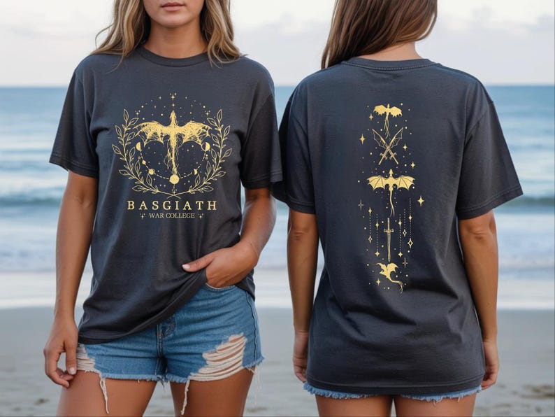 two women wearing black t-shirts with gold designs on them. The t-shirts have a dragon and star motif, and the text "BASGIATH WAR COLLEGE" is printed on them.