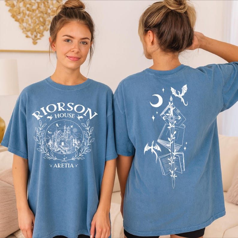 A woman wearing a blue t-shirt with a graphic design on the front and a back featuring a night scene with a castle, moon, and dragon.
