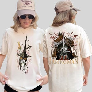 May include: Cream-colored t-shirt featuring a fantasy illustration on the front and back. The front design includes a helmet, sword, and floral elements. The back design features a warrior with a sword and the text "I AM NO MAN".