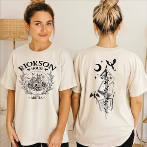 A woman wearing a white t-shirt with a graphic design on the front and a back featuring a black illustration of a castle, a crescent moon, and a dragon.