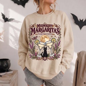 May include: A beige crewneck sweatshirt featuring a graphic print with the text "MIDNIGHT MARGARITAS" and "Moonlight, Magic & Sisterhood". The design includes two margarita glasses, a black cat, a full moon, candles, moths, and lavender.