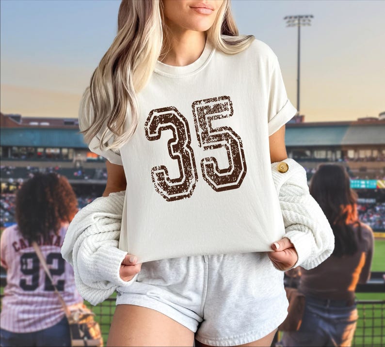 A woman wearing a white t-shirt with the number 35 on it, standing in front of a baseball stadium.