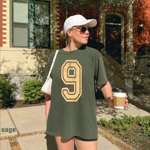 A woman wearing a white cap and a green t-shirt with the number 9 stands on a sidewalk, holding a coffee cup.
