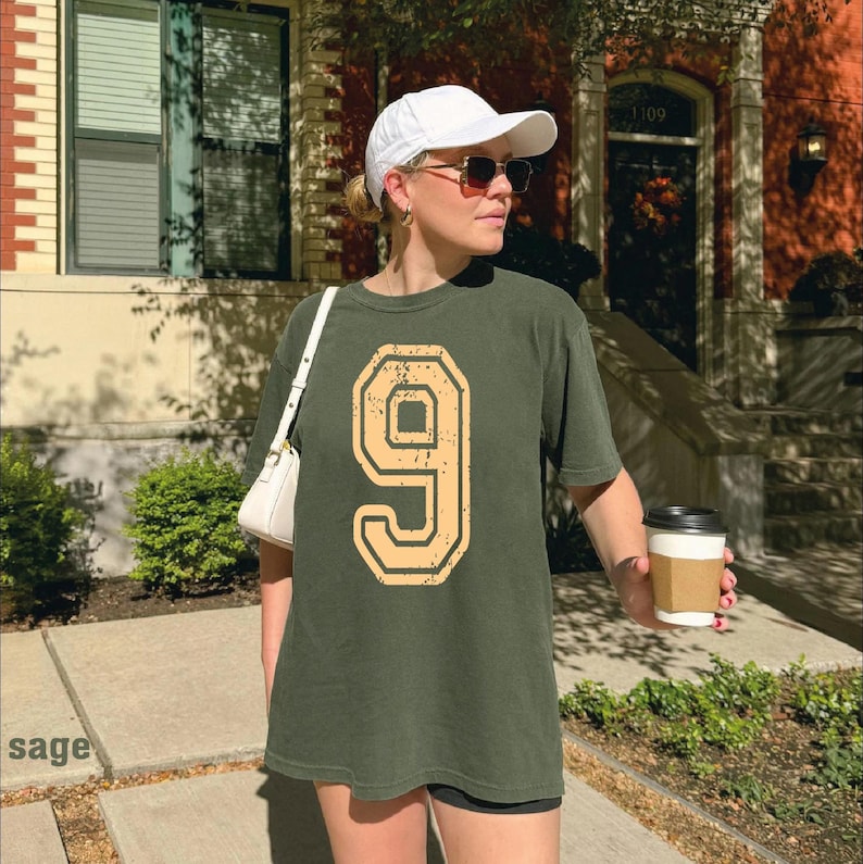 A woman wearing a white cap and a green t-shirt with the number 9 stands on a sidewalk, holding a coffee cup.