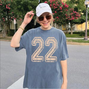 A woman wearing a white cap and sunglasses is standing on a street, smiling and posing for the camera.