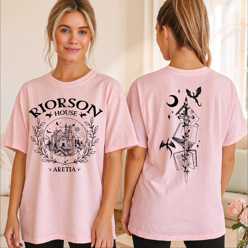 A woman wearing a pink t-shirt with a graphic design on the front and a back featuring a black illustration of a castle, a dragon, and a crescent moon.