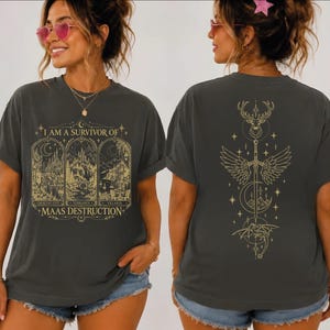 May include: A charcoal gray t-shirt featuring gold line art. The front displays three arched panels with landscapes labeled Crescent City, Terrasen, and Velaris, with the text I AM A SURVIVOR OF MAAS DESTRUCTION. The back shows a sword, wings, a stag, and a dragon.