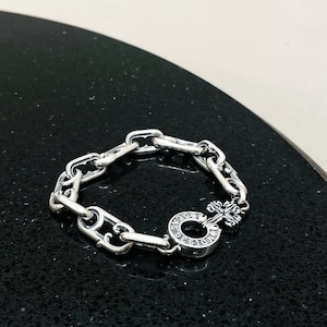 May include: A silver chain bracelet with a silver charm featuring a cross and the words "Chrome Hearts" in a circle.