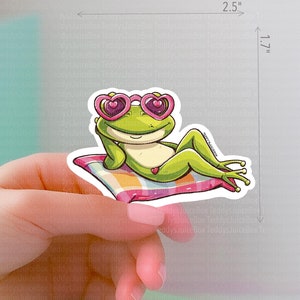 May include: A green frog wearing pink heart-shaped sunglasses is relaxing on a pink and white striped pillow. The sticker is 2.5 inches wide and 1.7 inches tall.