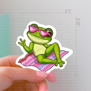 May include: A green frog wearing pink heart-shaped sunglasses is relaxing on a pink and white striped beach towel. The frog is sitting up with its legs stretched out and its arms resting on the towel. The frog is looking to the right.