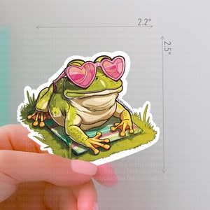 May include: A green frog wearing pink heart-shaped sunglasses is relaxing on a striped towel. The frog is surrounded by green grass and is looking directly at the viewer. The sticker is 2.2 inches wide and 2.5 inches tall.