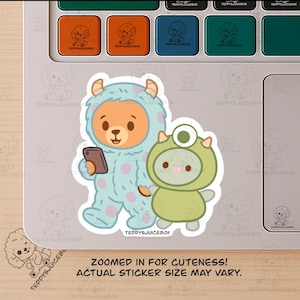 May include: A sticker featuring two cartoon characters. One is a bear in a blue monster costume holding a phone. The other is a green creature. The sticker has the text "ZOOMED IN FOR CUTENESS! ACTUAL STICKER SIZE MAY VARY."